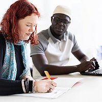 Zwei Studierende der ASH Berlin sitzen nebeneinander in einem Seminarraum. Eine Person schreibt etwas auf ein Papier, die andere Person schaut dabei zu.