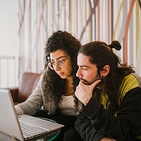 Zwei Personen sitzen auf einem Sofa, vor ihnen steht ein kleiner Tisch mit einem Laptop. Die Personen schauen auf den Laptop.