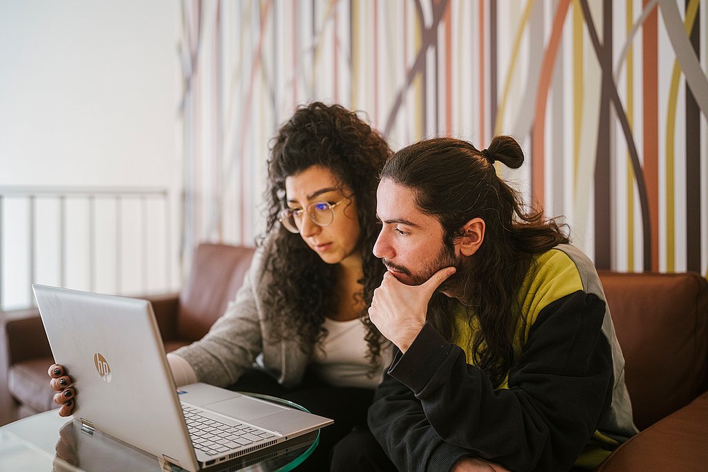 Zwei Personen sitzen auf einem Sofa, vor ihnen steht ein kleiner Tisch mit einem Laptop. Die Personen schauen auf den Laptop.