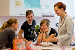 Drei Frauen sind in einer Werkstatt, haben verschiedene Werkzeuge in der Hand und vor sich auf dem Tisch liegen und diskutieren.