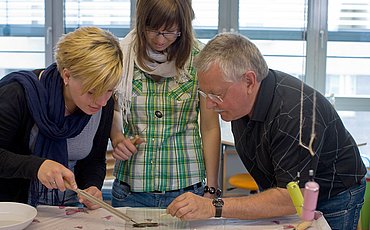 Zwei Studierende des pädagogischen Bachelor Studiengangs Erziehung und Bildung in der Kindheit beugen sich zusammen mit dem Leiter der Lernwerkstatt über eine Versuchsanordnung (Grashalm im Reagenzglas)