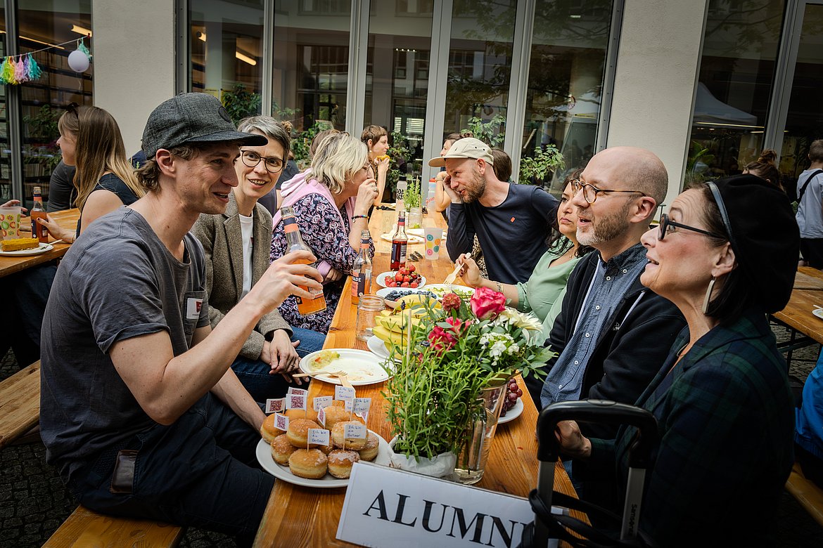 Eine Gruppe von Menschen sitzt um einen Tisch mit Obst und Gebäck. Alle lächeln und haben Spaß.