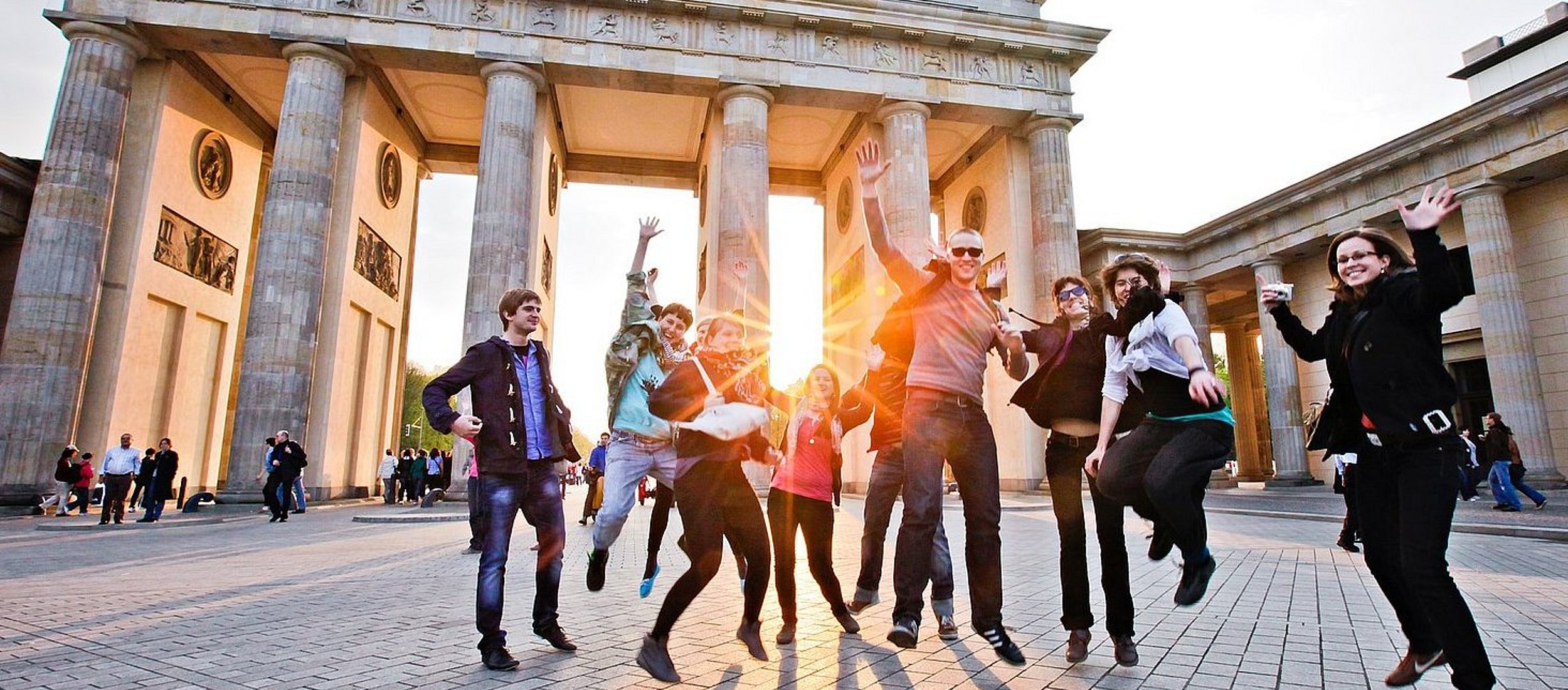 Vor dem Berliner Panorama des Brandenburger Tors springt eine Gruppe Studierender in die Luft.