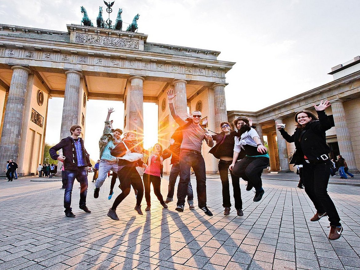 Vor dem Berliner Panorama des Brandenburger Tors springt eine Gruppe Studierender in die Luft.