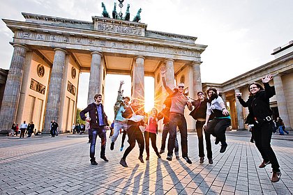 Vor dem Berliner Panorama des Brandenburger Tors springt eine Gruppe Studierender in die Luft.