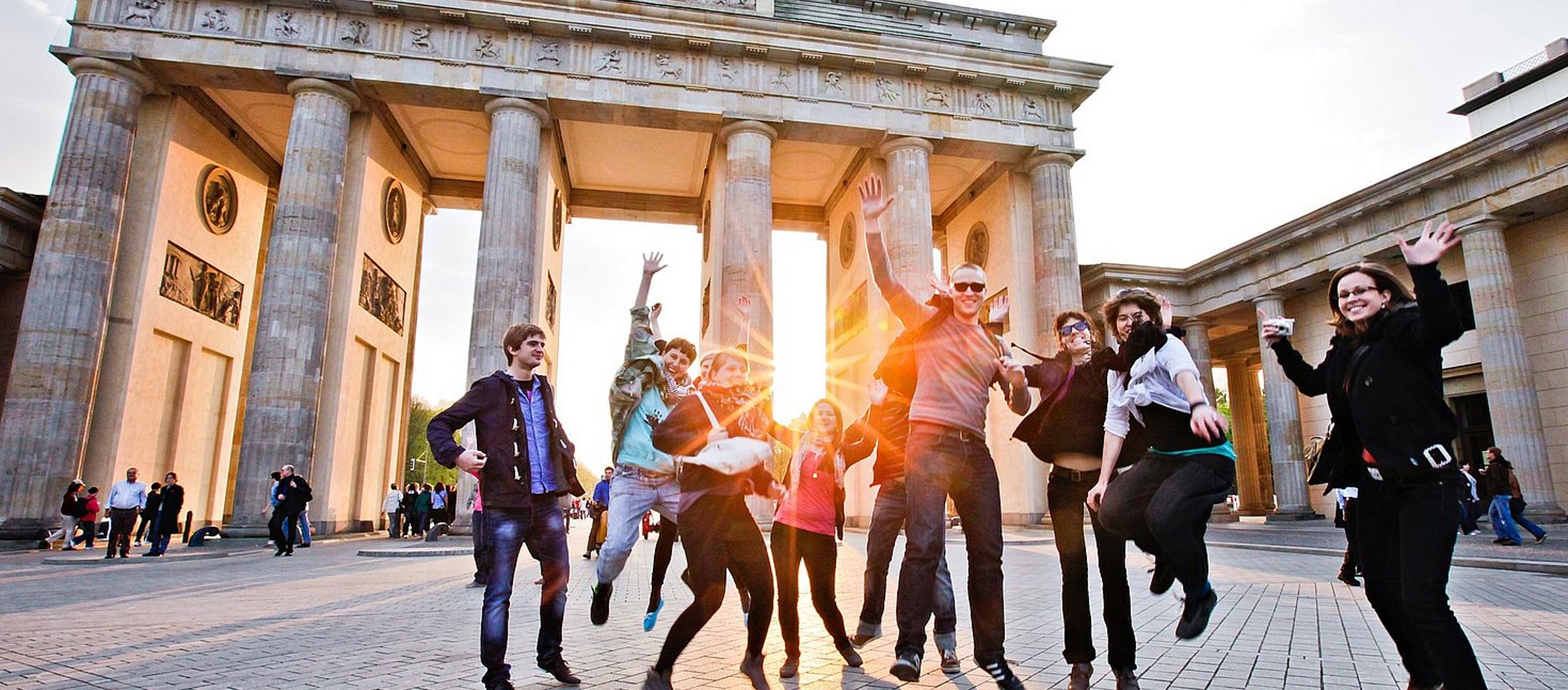 Vor dem Berliner Panorama des Brandenburger Tors springt eine Gruppe Studierender in die Luft.