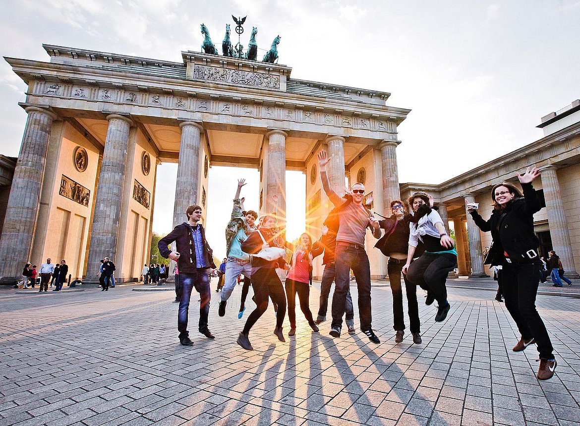 Vor dem Berliner Panorama des Brandenburger Tors springt eine Gruppe Studierender in die Luft.