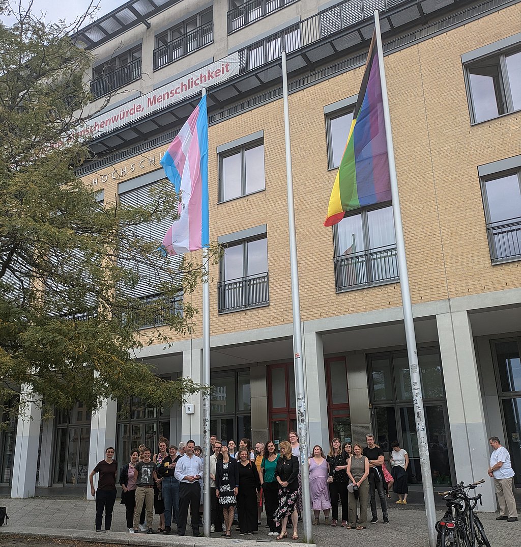 Auf dem Foto ist eine große Gruppe von Personen vor der ASH Berlin zu sehen, außerdem die Progressiveflag und die Transprideflag, die an den Fahnenmasten wehen. 