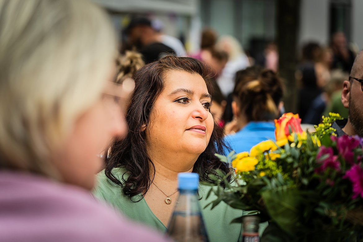 Blick auf eine Frau mit dunklem Haar und grüner Bluse. Sie sitzt an einem Tisch im Freien und wird im Vordergrund von Blumen und der Schulter einer Person eingerahmt.