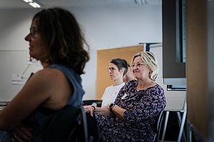 Drei Personen, die einem Vortrag aufmerksam folgen. Die Frau in der Mitte lächelt.