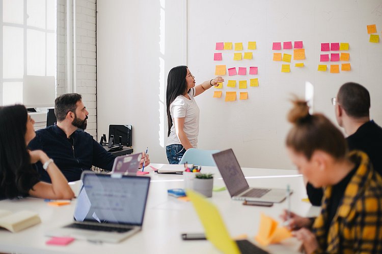 Menschen sitzen an einem Tisch und besprechen ein Thema. Auf dem Tisch stehen Laptops. Eine Person steht am Whiteboard. 