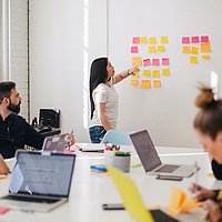 Menschen sitzen an einem Tisch und besprechen ein Thema. Auf dem Tisch stehen Laptops. Eine Person steht am Whiteboard. 