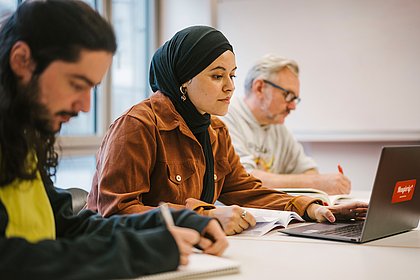 Auf dem Foto sind drei ASH-Studierende zu sehen, die an einem Tisch sitzen und arbeiten. Eine Person arbeitet am Laptop.