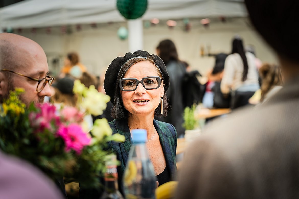 Blick auf eine Frau mit Brille, die lächelt und spricht. Sie sitzt an einem Tisch im Freien und wird im Vordergrund von Blumen und der Schulter einer Person eingerahmt.