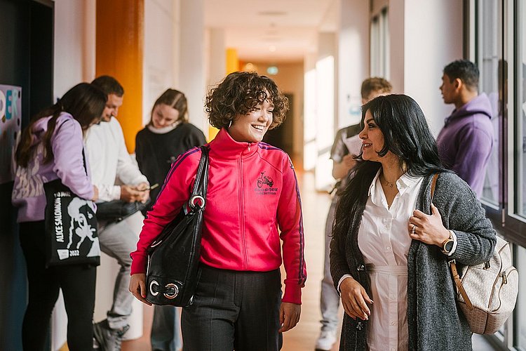 Auf dem Foto sind zwei ASH-Studierende zu sehen die einen Flur entlang laufen und sich dabei unterhalten. Im Hintergrund stehen weitere Personen, die sich ebenfalls unterhalten. 