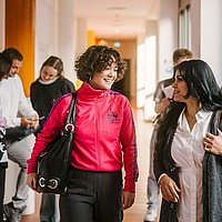 Auf dem Foto sind zwei ASH-Studierende zu sehen die einen Flur entlang laufen und sich dabei unterhalten. Im Hintergrund stehen weitere Personen, die sich ebenfalls unterhalten. 