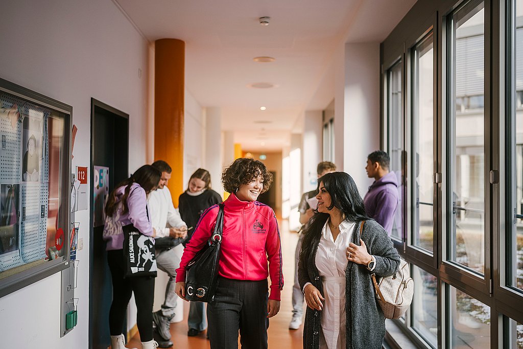 Auf dem Foto sind zwei ASH-Studierende zu sehen die einen Flur entlang laufen und sich dabei unterhalten. Im Hintergrund stehen weitere Personen, die sich ebenfalls unterhalten. 