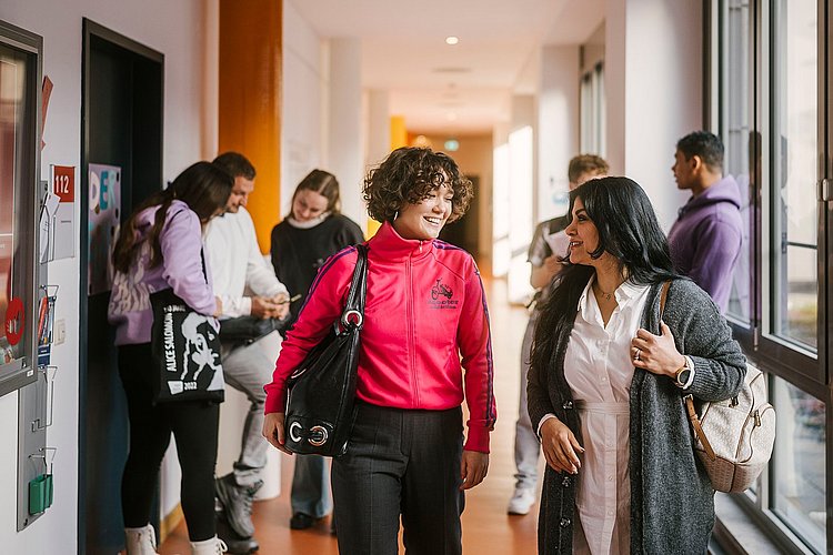 Auf dem Foto sind zwei ASH-Studierende zu sehen die einen Flur entlang laufen und sich dabei unterhalten. Im Hintergrund stehen weitere Personen, die sich ebenfalls unterhalten. 