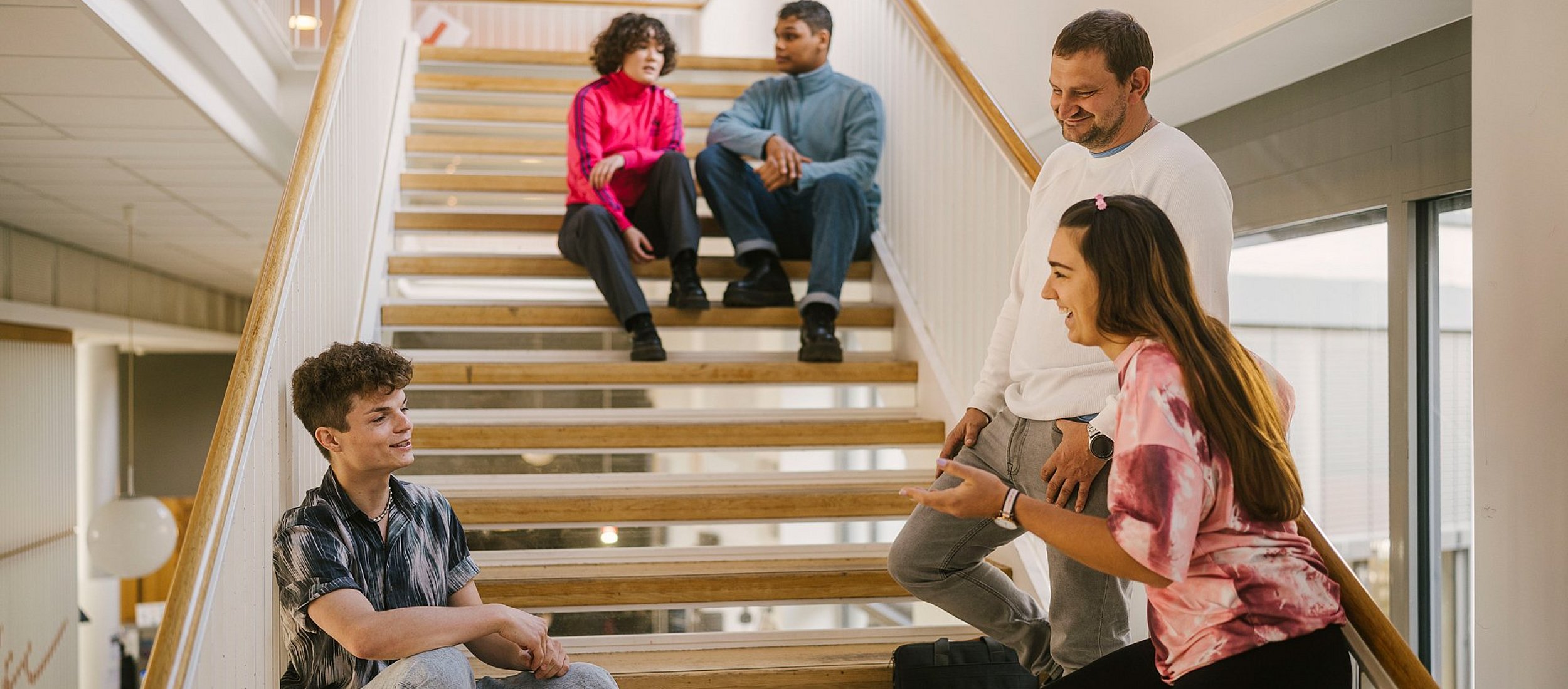 Auf dem Foto sind Studierende der ASH Berlin zu sehen, die auf einer Treppe in der Hochschule stehen oder sitzen und sich miteinander unterhalten.