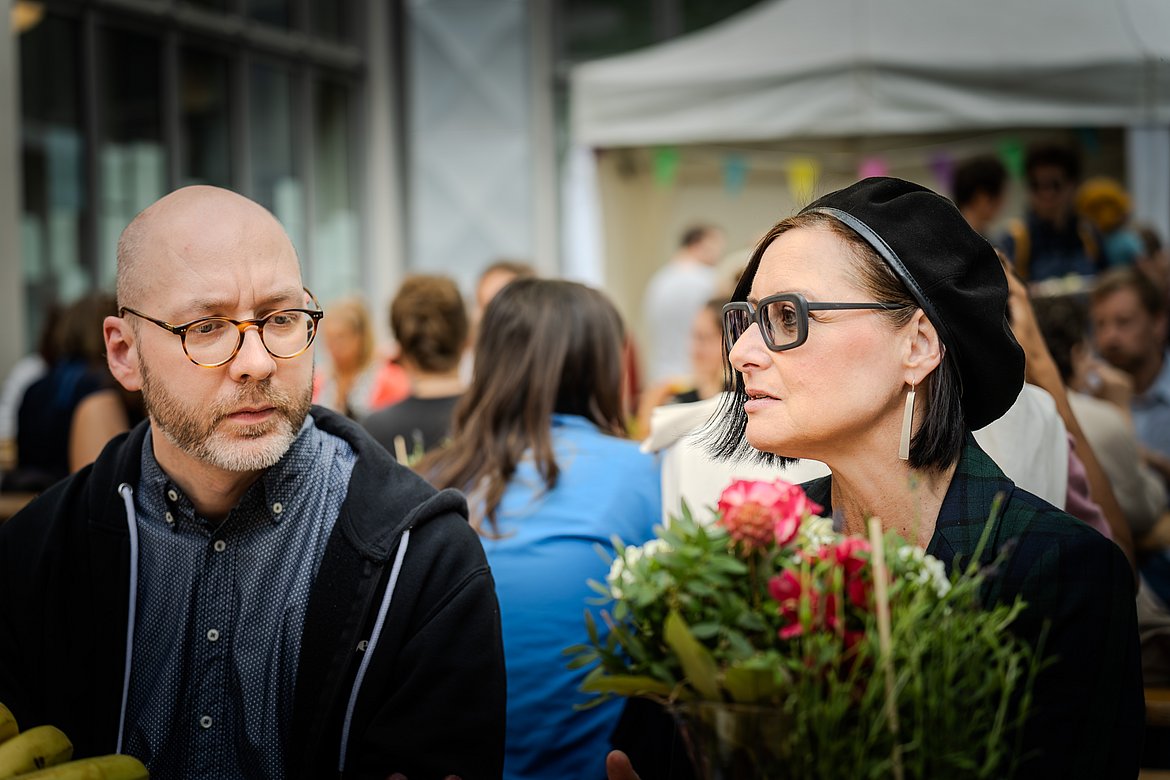 Eine Frau mit Brille spricht mit einem Mann, der ebenfalls eine Brille trägt.