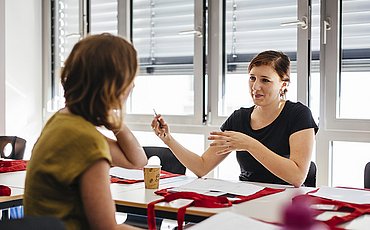 Zwei Studentinnen sitzen sich an einem Tisch gegenüber. Eine Studentin erklärt der anderen etwas und gestikuliert mit ihren Armen.