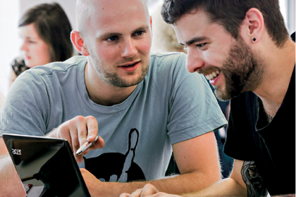 Zwei Studenten sitzen vor einem Laptop und scheinen sich zu amüsieren