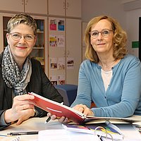 Corinna Schmude und Anja Voss sitzen an einem Besprechungstisch und halten das Berliner Bildungsprogramm für Kitas und Kindertagespflege in den Händen