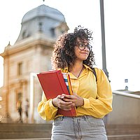 Auf dem Foto ist eine Frau zu sehen, die eine gelbe Jacke trägt und ein rotes Buch in der Hand hält. Sie steht auf einer Treppe vor einem historischen Gebäude und schaut lächelnd nach rechts. 