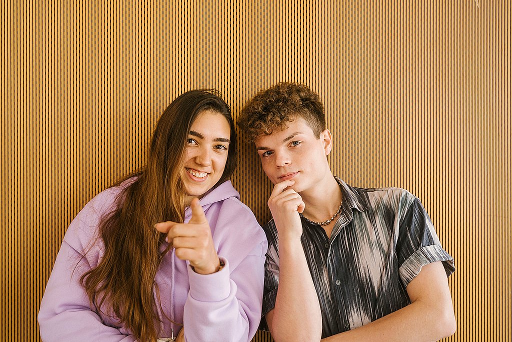 Two people stand in front of a monochrome wall and look directly into the camera, smiling.