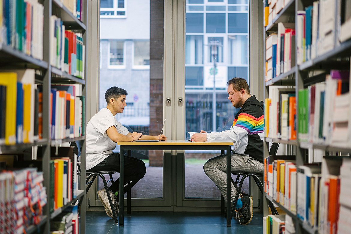 Zwei Studierende der ASH Berlin sitzen an einem Tisch in der Bibliothek und arbeiten.