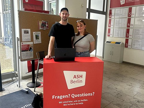 Zwei Studentische Mitarbeiter_innen von dem Projekt "Start with a PEER" stehen an dem Infodesk für die Vermittlung.