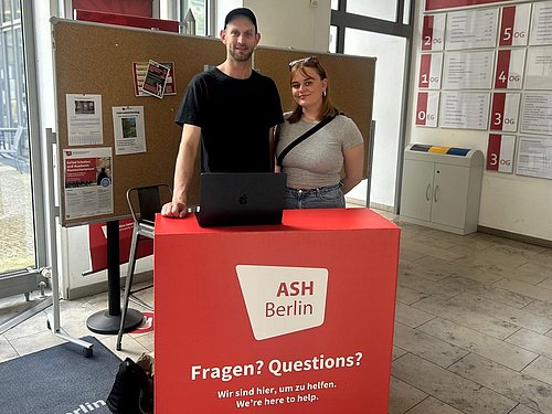 Zwei Studentische Mitarbeiter_innen von dem Projekt "Start with a PEER" stehen an dem Infodesk für die Vermittlung.