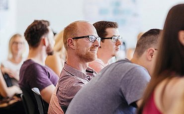 Lehrsituation des Master of Science Management und Qualitätsentwicklung im Gesundheitswesen Eine Reihe Studierender im Unterricht Fokus auf einen Studenten mit Brille der lächelnd zuhört