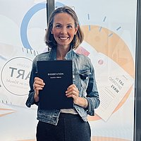 Auf dem Foto ist Jennifer Hübner zu sehen. Sie steht in einem Copyshop und hält ihre frisch gedruckte Dissertation in den Händen. 