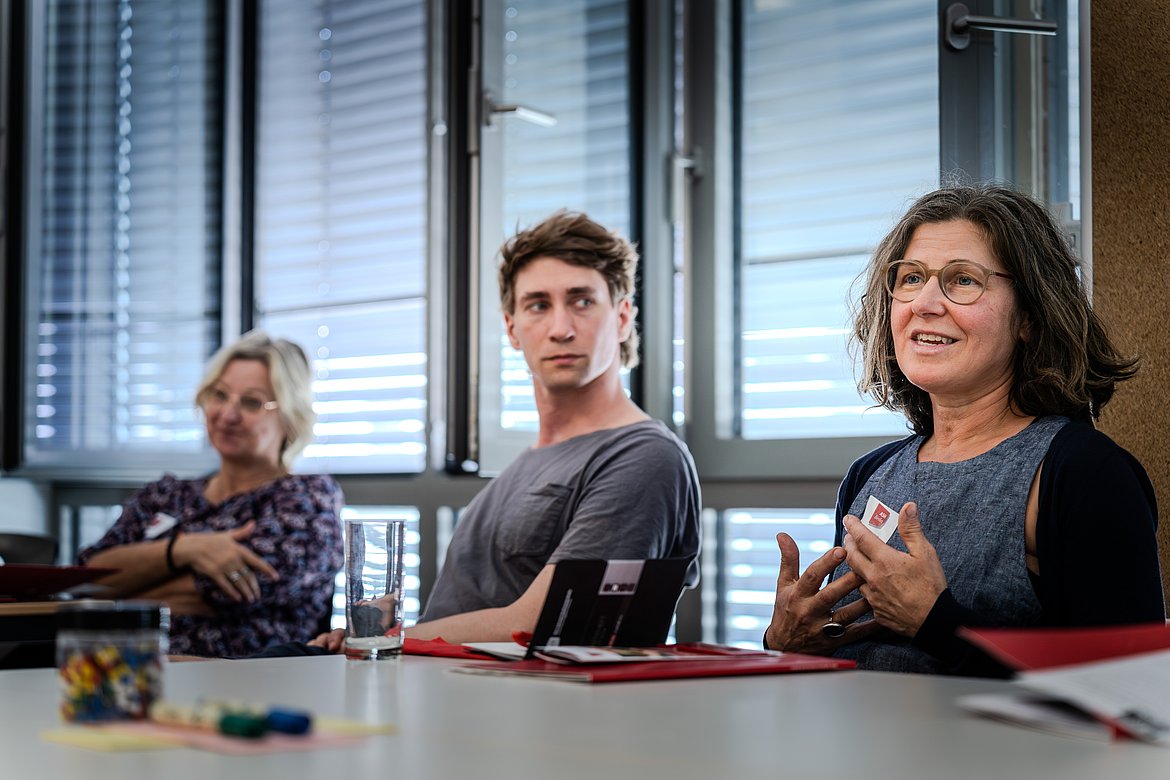 Drei Personen sitzen während eines Workshops an einem Tisch. Die Person auf der rechten Seite spricht, und die beiden anderen hören ihr zu.